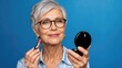 © Andrii  - Stylish Senior Woman Applying Lipstick While Looking in Compact Mirror Against Blue Background