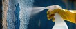 © Viktor - Hand in yellow rubber glove uses spray bottle to clean blue wall. Person applies liquid detergent on textured surface to remove dirt stain. Worker performs home maintenance renovation task using