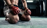 Fighter kneeling, sweating profusely with open-finger gloves on gym mat, representing intense training and physical exhaustion.