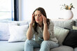© Daniel - Woman feeling intense migraine pain, holding her temples while sitting on a comfortable couch in a bright living room, reflecting discomfort and health issues