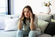 © Daniel - Woman sitting on a sofa, eyes closed and hands on her temples, suffering from a severe headache, expressing discomfort and pain, a common symptom of stress, burnout, or fatigue
