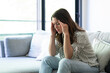 © Daniel - Woman seated on sofa in bright modern living room, pressing hands to temples in pain and discomfort from a severe headache or migraine, looking exhausted and stressed
