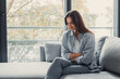 © Daniel - Young woman feeling intense abdominal pain and discomfort, sitting on a sofa at home and holding her stomach with both hands, experiencing a health issue