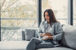 © Daniel - Woman sitting on a sofa in a modern apartment, experiencing discomfort and holding her stomach due to pain, possibly from indigestion, illness, or period cramps, needing relief and medical care