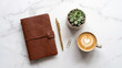 © Ali - Brown leather journal placed on white marble surface alongside golden pen succulent plant and ceramic cup of hot coffee