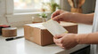 © ViskBx - Person holding a blank shipping label above a cardboard box on a kitchen counter, ready to prepare a package for delivery