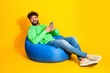 © deagreez - Smiling young man in green hoodie relaxing on blue beanbag while using smartphone against bright yellow background