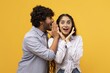 © Prostock-studio - Excited Young South Asian Couple Smiling And Whispering Secrets To Each Other Against A Bright Yellow Background, Showing Joyful Emotion In A Fun And Playful Indoor Setting. Lifestyle Concept.