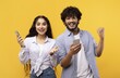 © Prostock-studio - Excited Young Couple, Hispanic And Asian, Celebrating A Successful Event While Holding Smartphones In A Studio Setting With Yellow Background, Displaying Happiness And Joyful Emotion.
