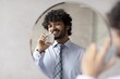 © Prostock-studio - Smiling Young South Asian Man Holding Bottle Of Perfume And Applying It In Front Of Mirror In Modern Office Setting, Representing Personal Care And Professional Lifestyle. Business Concept.