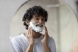 © Prostock-studio - Smiling Young Man Of South Asian Ethnicity Applying Shaving Cream While Standing In Front Of A Mirror At Home, Engaging In Personal Grooming For A Fresh Look In His Daily Routine.