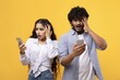 © Prostock-studio - Worried South Asian Young Adult Male And Female Looking At Smartphone With Shocked Expressions, Standing Together In Studio Setting. Emotion Of Surprise Presenting Digital Communication Concept.