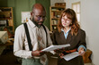 © AnnaStills - Middle aged African American man and young adult Caucasian woman reviewing documents in retro office. Image suitable for vintage corporate communication and business history campaigns
