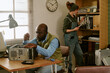 © AnnaStills - Middle aged African American man repairing computer hardware while Caucasian woman organizes files in retro office. Image suitable for vintage Information Technology support and tech history concepts