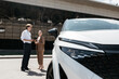 © wedmoments.stock - Man presenting a modern white car to a woman near a dealership during a consultation before purchase.