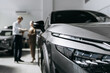 © wedmoments.stock - Front section of a modern car in close-up inside a dealership while customers receive consultation in the background.