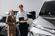 © wedmoments.stock - Man consulting a woman about a new car beside the vehicle in a modern dealership.