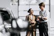 © wedmoments.stock - Man with a tablet consulting a woman beside a new car in a modern dealership.