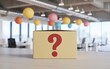 © Lamia - A cardboard box with a large red question mark printed on the front sitting on a wooden desk in a blurred modern office environment with colorful hanging paper lanterns