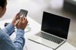 © MINAE - Person using smartphone and pen while working at a desk with a laptop, managing communication, planning tasks, and multitasking in a productive office environment