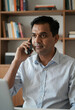 © Pixel-Shot - Business communication and remote work, indian man talking on smartphone in home office with bookshelf background