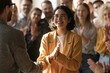 © Ravi - Happy woman wearing yellow blazer shaking hands with colleague while coworkers applaud