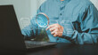 © Krittamet - Audit, compliance checking, administrative review, verification, governance, and risk control. Professional using a magnifying glass to inspect digital documents on a laptop.