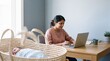 © AdPremium - Indian mother working on laptop at home while newborn baby sleeps in a woven bassinet nearby