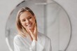 © Prostock-studio - Happy Young Woman Smiling While Gently Applying Skincare Cream To Her Face In Front Of A Bathroom Mirror, Enjoying Self-care Routine At Home, Creating A Relaxing Spa Environment.