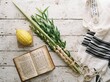 © AI img - Traditional Jewish Sukkot holiday symbols featuring a lulav with etrog, an open prayer book, and a tallit shawl displayed on a rustic white wooden background, top view.