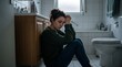 © olga - A woman sitting on the floor of a bathroom, looking down with her head in her hands, appearing distressed and upset