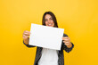 © Daniel - Young woman smiling and holding a blank white paper sign against a vibrant yellow background, ideal for advertising, promotions, announcements or custom text and marketing use