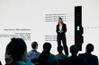 © DC Studio - Young adult presents a mobile phone announcement on stage with promotional visuals, advertising a new release and investment deal during a business convention and product launch event for investors.