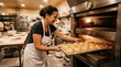 © Healy - Smiling female baker in white apron sliding a metal baking sheet filled with raw sesame-seed burger buns into a glowing commercial deck oven in a busy artisan bakery kitchen