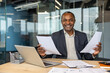 © Liubomir - Experienced businessman reviews documents at his desk, smiling confidently beside a laptop and paperwork in a modern corporate office, conveying professional success and satisfaction