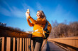 © 5ph - Tourist woman making selfie picture in autumn forest on wooden path - travel blogging. Female hiker recording video content for social media. Modern technology and networking concept.