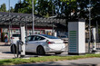 © K - A white electric car charges at a public fast-charging station, the roadside infrastructure that decides how quickly a country can retire its petrol fleet.