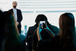 © DC Studio - Young woman takes photos at a business convention to post online and build career experience, while the conference summit in an auditorium provides social media content and professional growth.