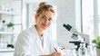 © olga_demina - A female scientist in a white coat writes in a laboratory, smiling at the camera with a compound light microscope on the table nearby