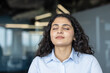 © Liubomir - Young woman closing eyes, finding calm and peace through deep breathing and mindfulness in a corporate office setting, promoting stress relief and mental well-being in the workplace