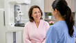 © Artyom - Older woman in a pink robe smiles while speaking with a healthcare professional beside a mammography machine during a breast screening appointment