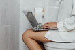 © Ratirat - Woman using laptop while sitting on toilet in bathroom, combining remote work, technology, multitasking, restroom habits, productivity, work from home routine. Rushing to get work done.