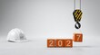 © Badhon - Industrial hoist hook lifting orange wooden block with number seven to complete year 2027 next to white hard hat on clean gray studio background.