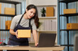 © Natee Meepian - Asian woman small business owner checking laptop while holding parcel box