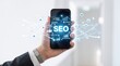 © Emma - Hand holding smartphone displaying SEO search engine optimization with floating holographic icons for traffic growth, visibility, keywords, analytics and marketing strategy