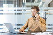 © Liubomir - Businessman concentrating on a phone call while simultaneously working on a laptop at a modern office desk, demonstrating multitasking and professional communication in a corporate environment