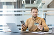© Liubomir - Stressed man looking at the camera with a surprised expression, holding a smartphone and credit card, experiencing online payment problems while sitting at his office desk with a laptop