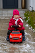 © sommersby - Happy little toddler girl in warm red puffer jacket and pink knit beanie playing on red ride-on toy car outside on cold winter day.