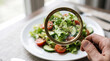 © Kristelinmind - Woman hand examining fresh vegetable salad through magnifying glass. Concept of orthorexia and obsessive control of food intake. Disordered eating and unhealthy relationship with nutrition.