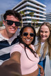 © CarlosBarquero - Vertical photo of three diverse young friends smiling together for a selfie in a sunny modern urban setting, conveying friendship, fun and connection.